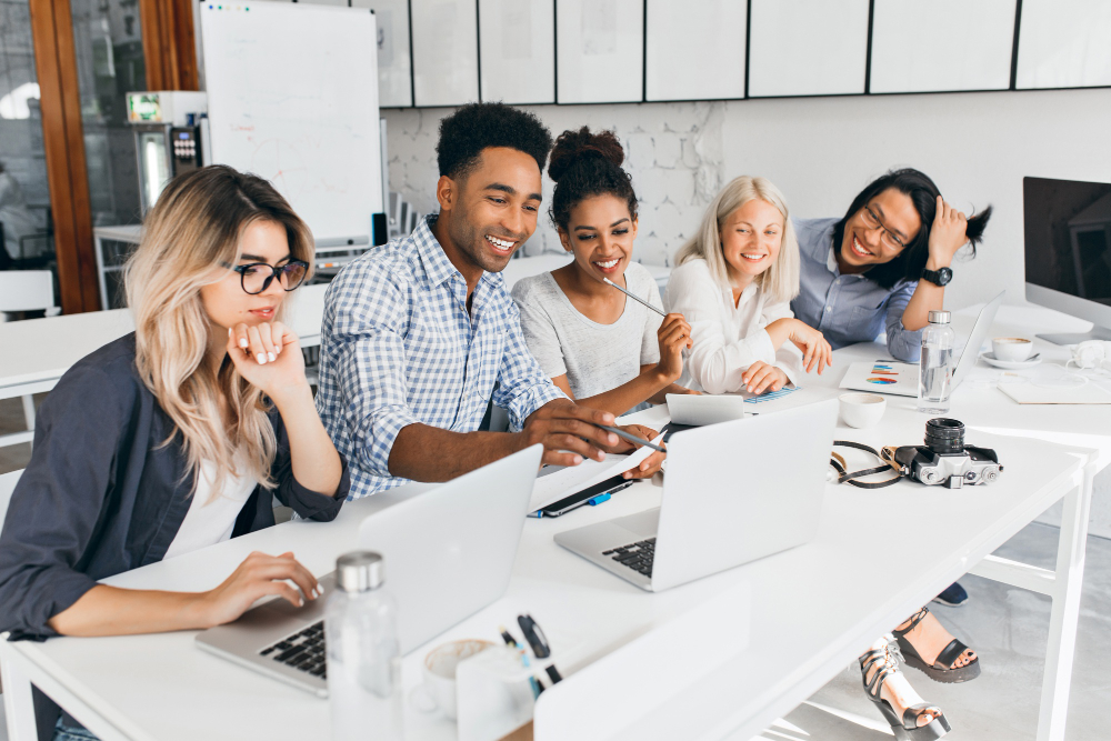 smiling-african-student-pointing-with-pencil-laptop-screen-concentrated-blonde-woman-glasses-propping-chin-with-hand-while-working-with-computer-office (1)